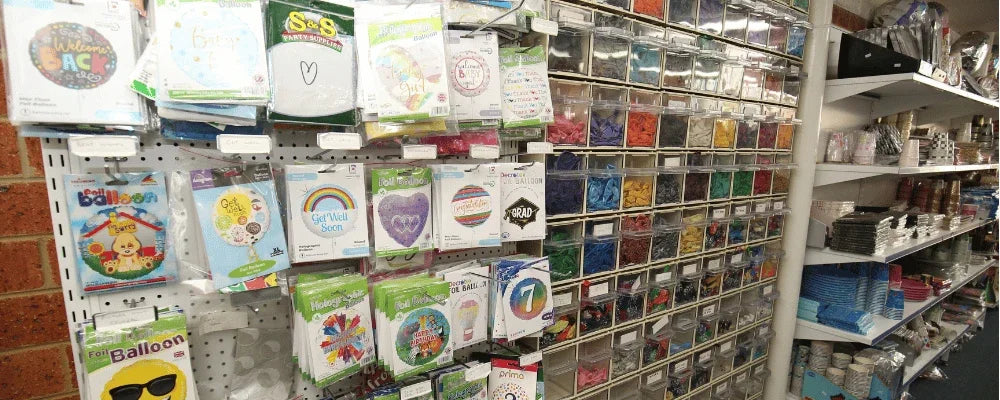 Retail display of various foil balloons and colorful party supplies in storage bins.